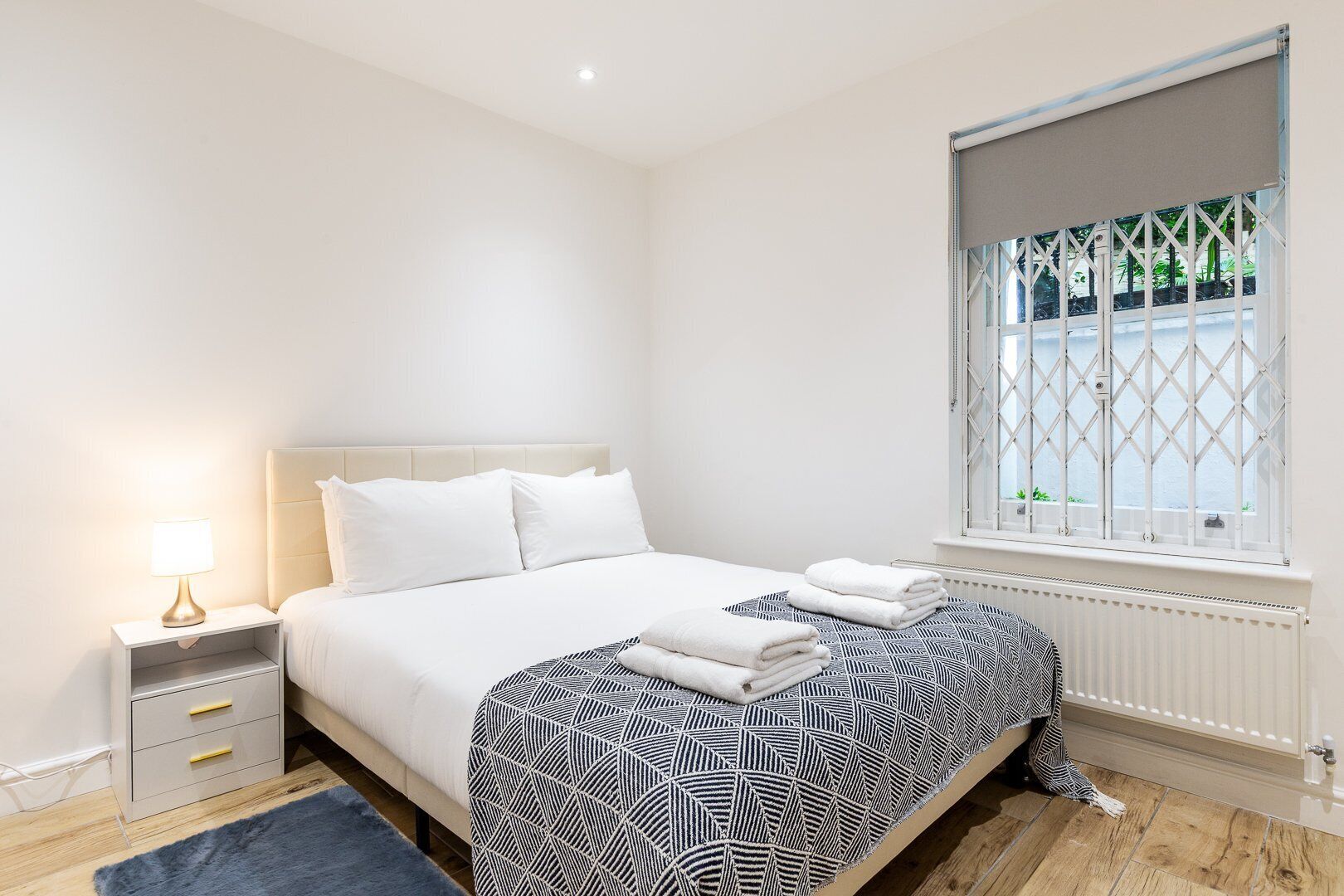 Quarto com cama, mesa de cabeceira, luminária, janela e radiador.