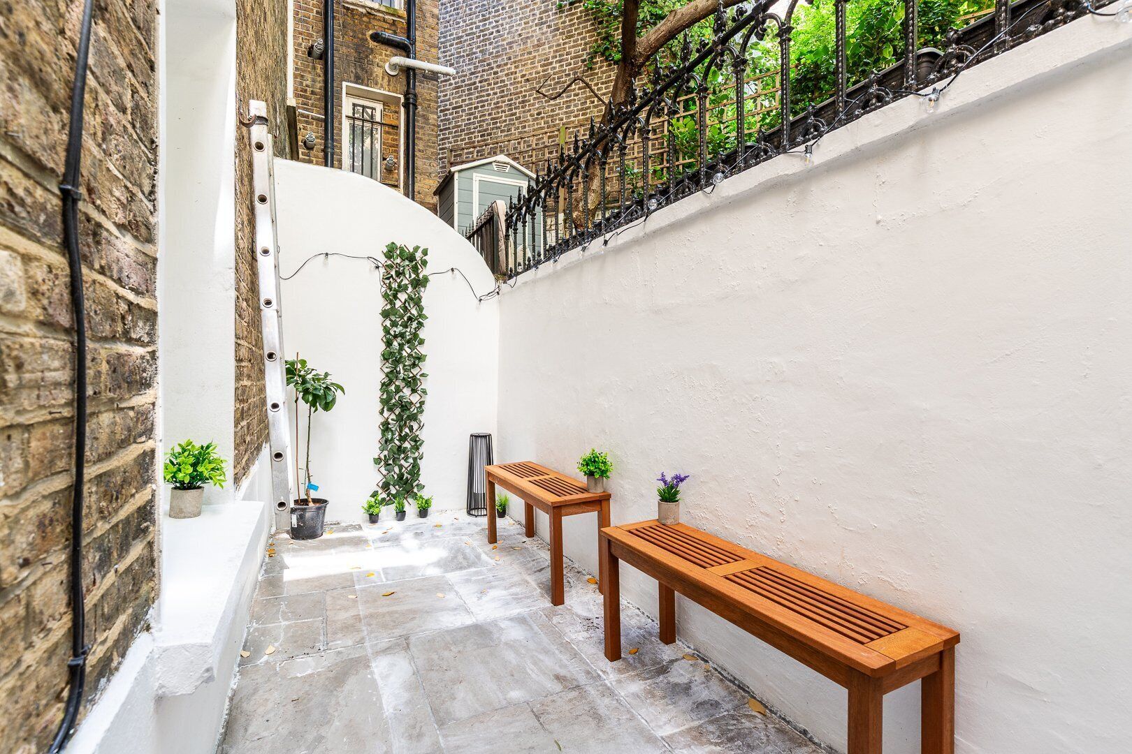 Une ruelle étroite bordée d’un mur blanc, d’un mur de briques et d’une clôture en métal. On y trouve deux bancs en bois et plusieurs plantes en pot.