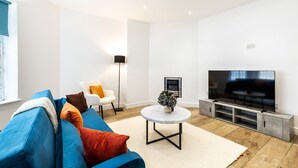Ett modernt vardagsrum med en blå soffa, en vit fåtölj, ett runt soffbord, en tv och en öppen spis.