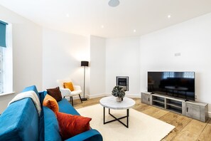 A modern living room with a blue sofa, a white armchair, a round coffee table, a TV, and a fireplace. - Stylish Holland Park Patio Apartment (London)