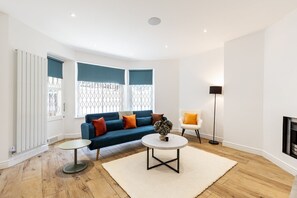 A living room with a blue sofa, two side tables, a white rug, and a floor lamp. - Stylish Holland Park Patio Apartment (London)