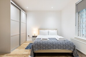 A bedroom with a bed, bedside table, lamp, and a window with a view. - Stylish Holland Park Patio Apartment (London)