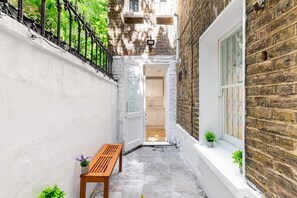 A narrow alleyway with a wooden bench, a potted plant, and a door leading to a room with a window. - Stylish Holland Park Patio Apartment (London)