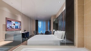 Ein modernes Hotelzimmer mit einem großen Bett, einem Schreibtisch mit Stuhl, einem an der Wand montierten Fernseher und einem großen Fenster mit Vorhängen.