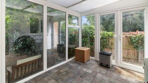 A sunroom with large glass doors, a wooden bench, a woven stool, and a trash bin. - Peaceful Cambridge haven - walk to Addenbrookes (Cambridgeshire)
