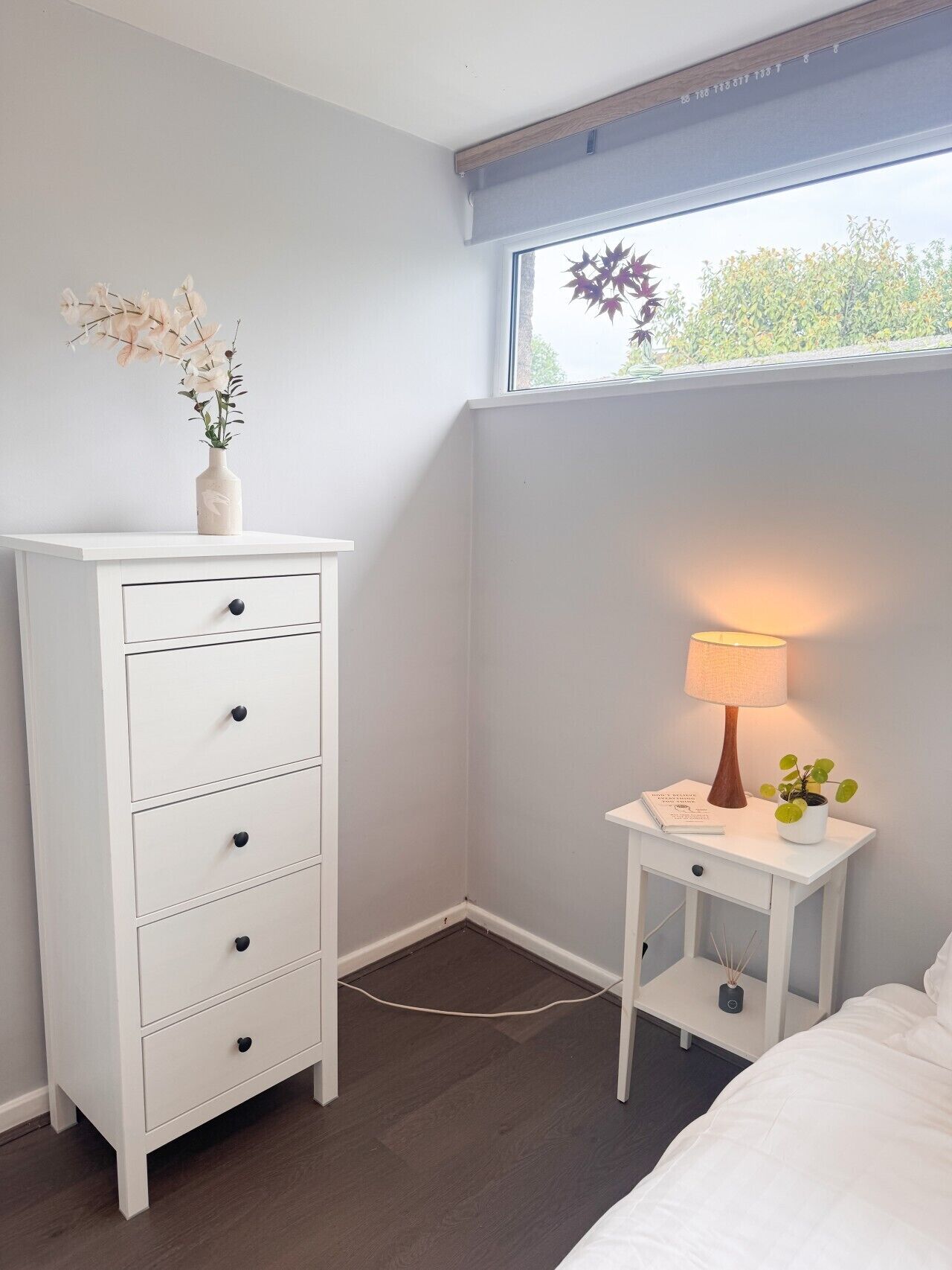 A bedroom with a white dresser, a bedside table with a lamp, and a window with a view of trees.
