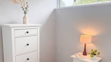 A bedroom with a white dresser, a bedside table with a lamp, and a window with a view of trees.