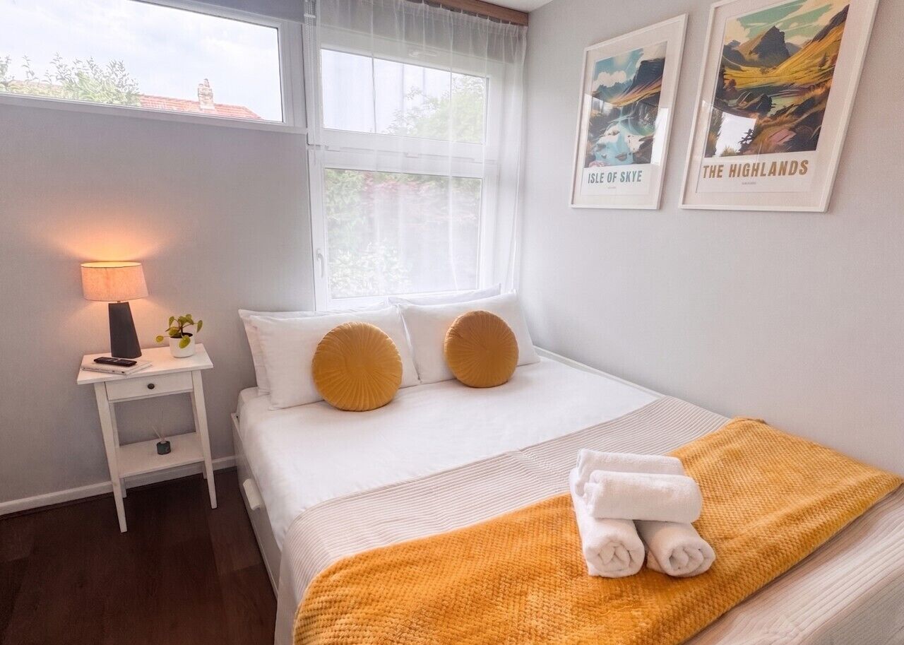A neatly made bed with white bedding and two mustard-colored pillows, a bedside table with a lamp, and two framed pictures on the wall.