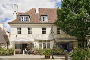 Un edificio de dos plantas con un letrero que dice 'The Somers Town Coffee House'. Hay áreas de estar al aire libre con mesas y sillas, y se ven macetas con plantas.