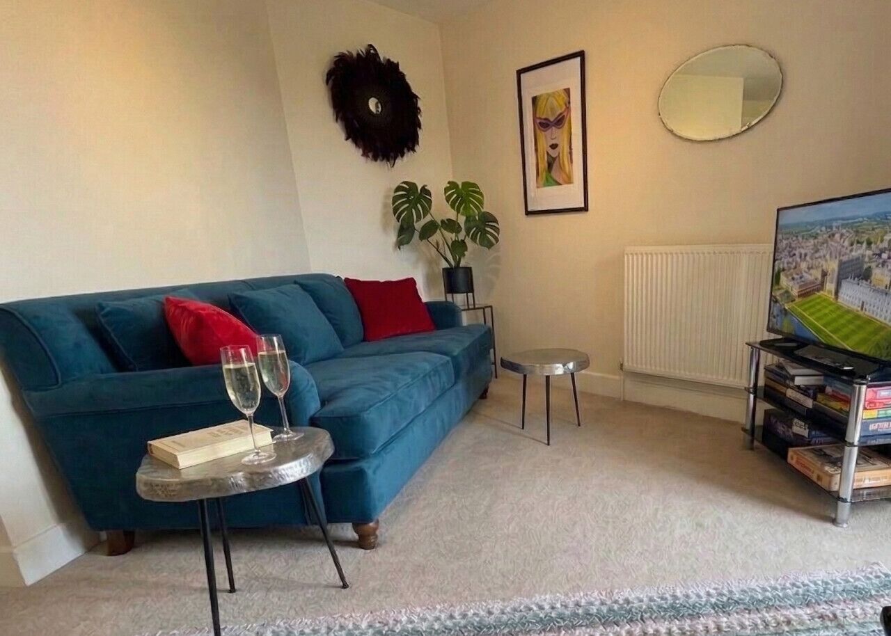 A living room with a blue sofa, red cushions, a small round table with glasses, a potted plant, a framed picture, a mirror, and a bookshelf with books.