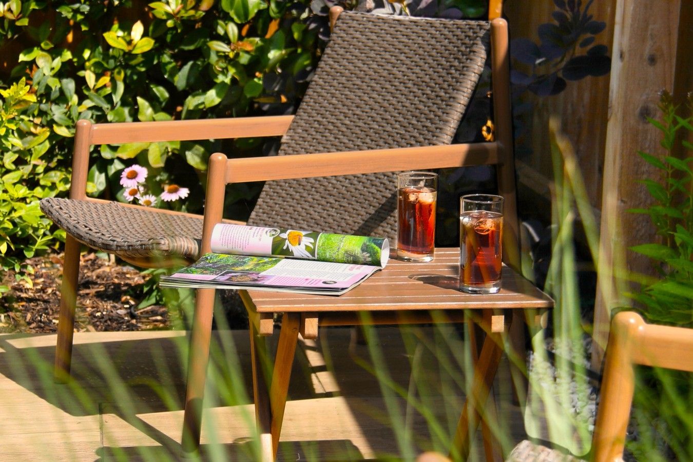 A wooden chair with a woven backrest and seat, a table with a magazine and two glasses of iced tea, and a garden with green foliage and flowers in the background.