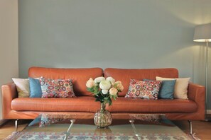 Una habitación con un sofá apoyado en una pared, un florero con flores sobre una mesa y una lámpara.
