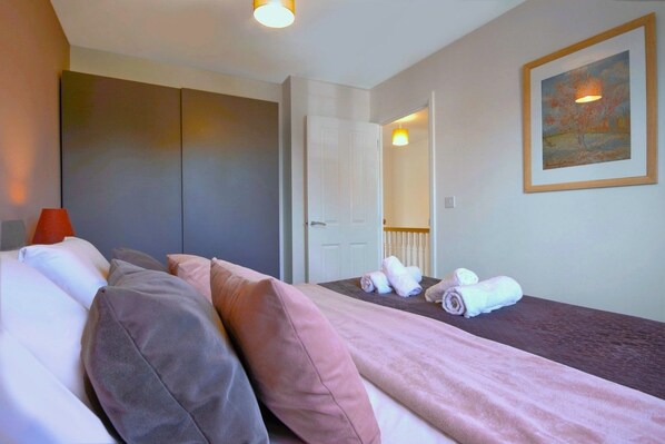A hotel room with a bed, pillows, a bedside table, a lamp, a painting on the wall, and a door leading to another room.