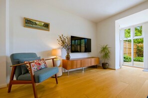 Een woonkamer met een bank, een bijzettafeltje met een lamp, een aan de muur bevestigde televisie en een potplant.