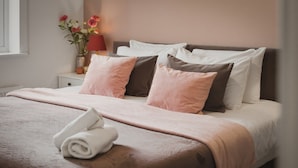 Uma cama bem arrumada com cores rosa e branco, um vaso com flores e uma luminária em uma mesa de cabeceira.