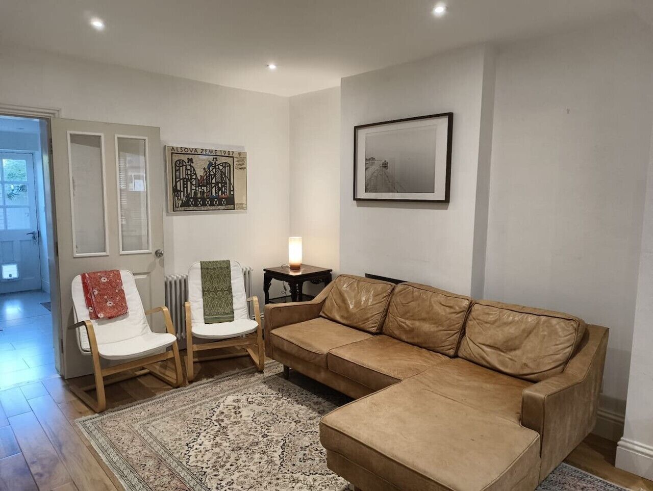 A living room with a beige sofa, two white chairs, a patterned rug, a framed picture, and a radiator.