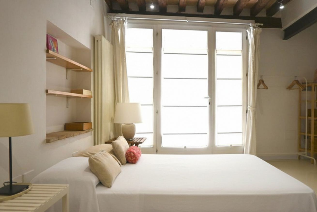 A bedroom with a bed, a bedside table, a lamp, a window with curtains, and a wardrobe.