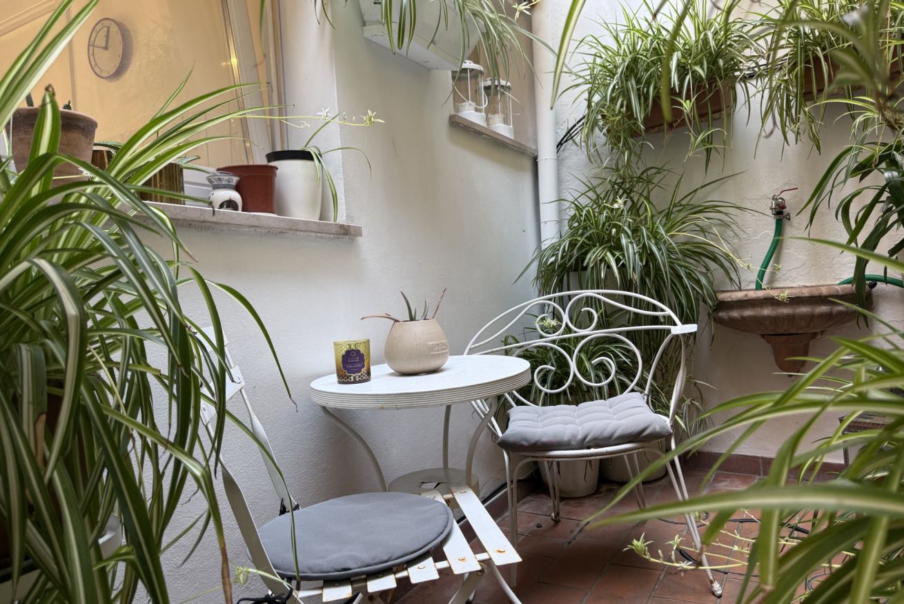 Un pequeño patio con una silla y mesa de metal blanco, rodeado de plantas en macetas y un muro blanco.