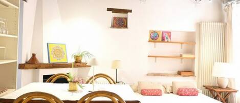 A room with a dining table, chairs, a bed, a shelf with books, and a small table with a lamp.