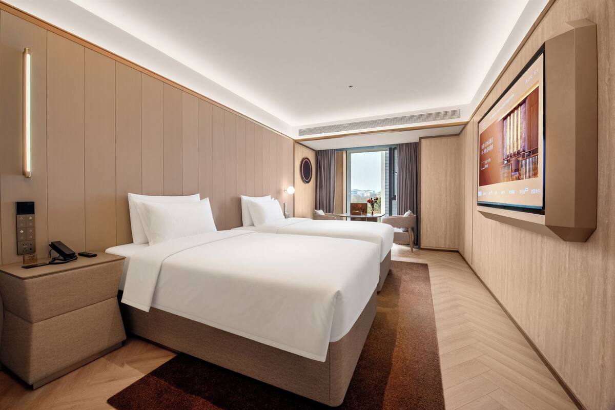 Ein Hotelzimmer mit zwei Betten, einem an der Wand befestigten Fernseher, einem Schreibtisch und einem Nachttisch.