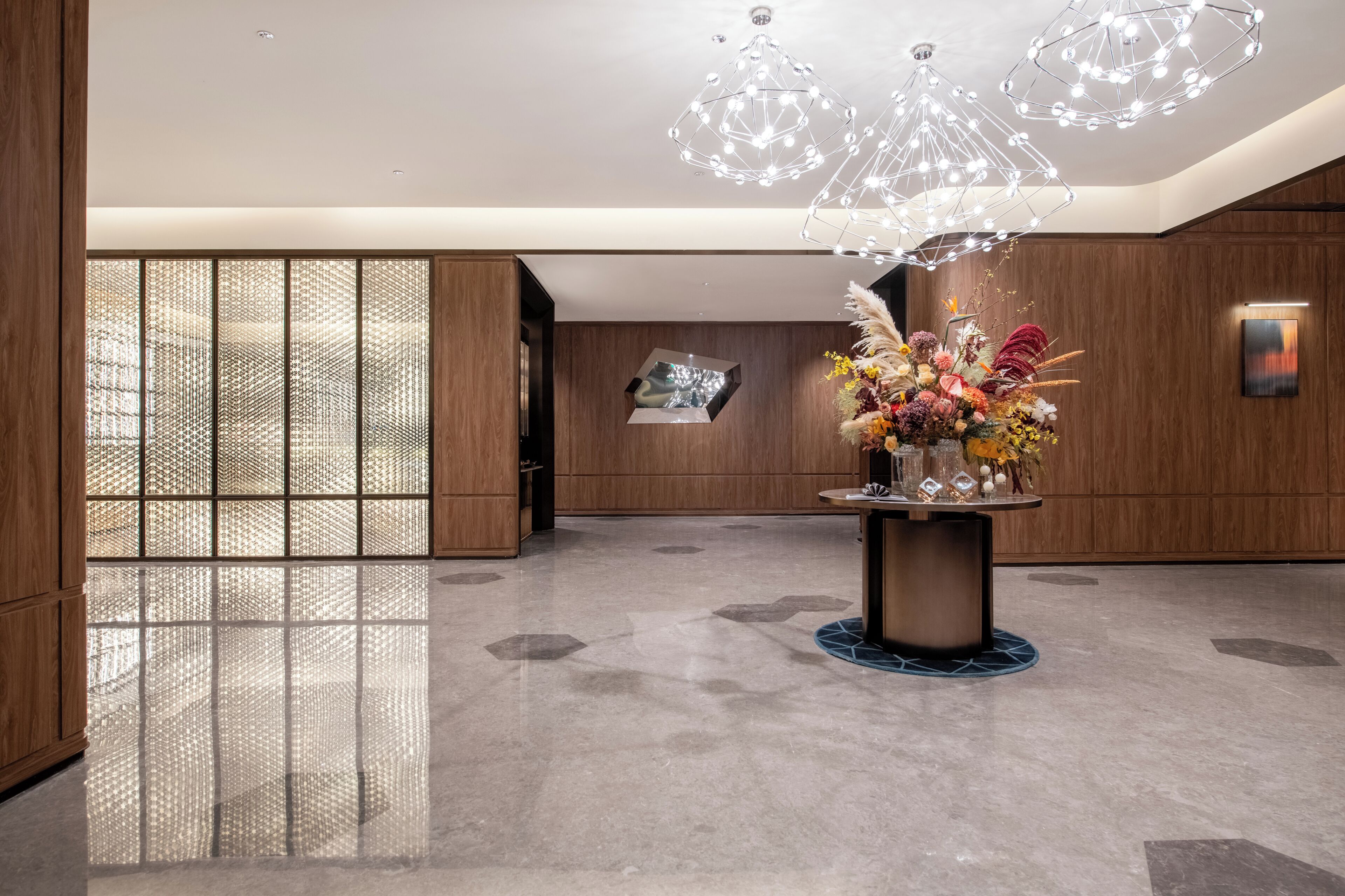 A modern lobby with a polished floor, a central table with a floral arrangement, and geometric light fixtures.
