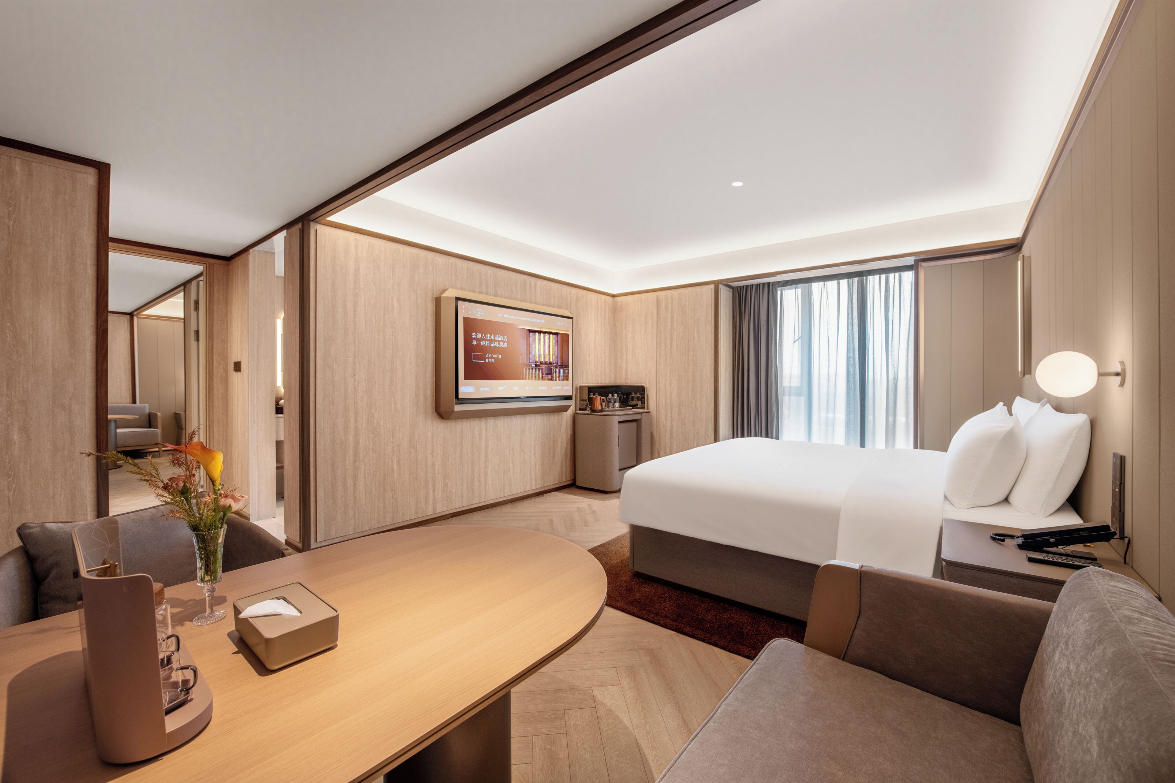 A hotel room with a bed, a desk, a chair, a small table, a vase with flowers, a framed picture on the wall, and a window with curtains.