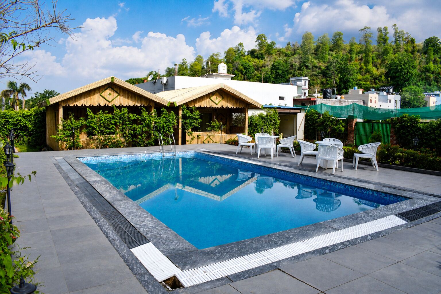 Una piscina con borde de baldosas, rodeada de sillas blancas y una zona de estar pequeña y cubierta.