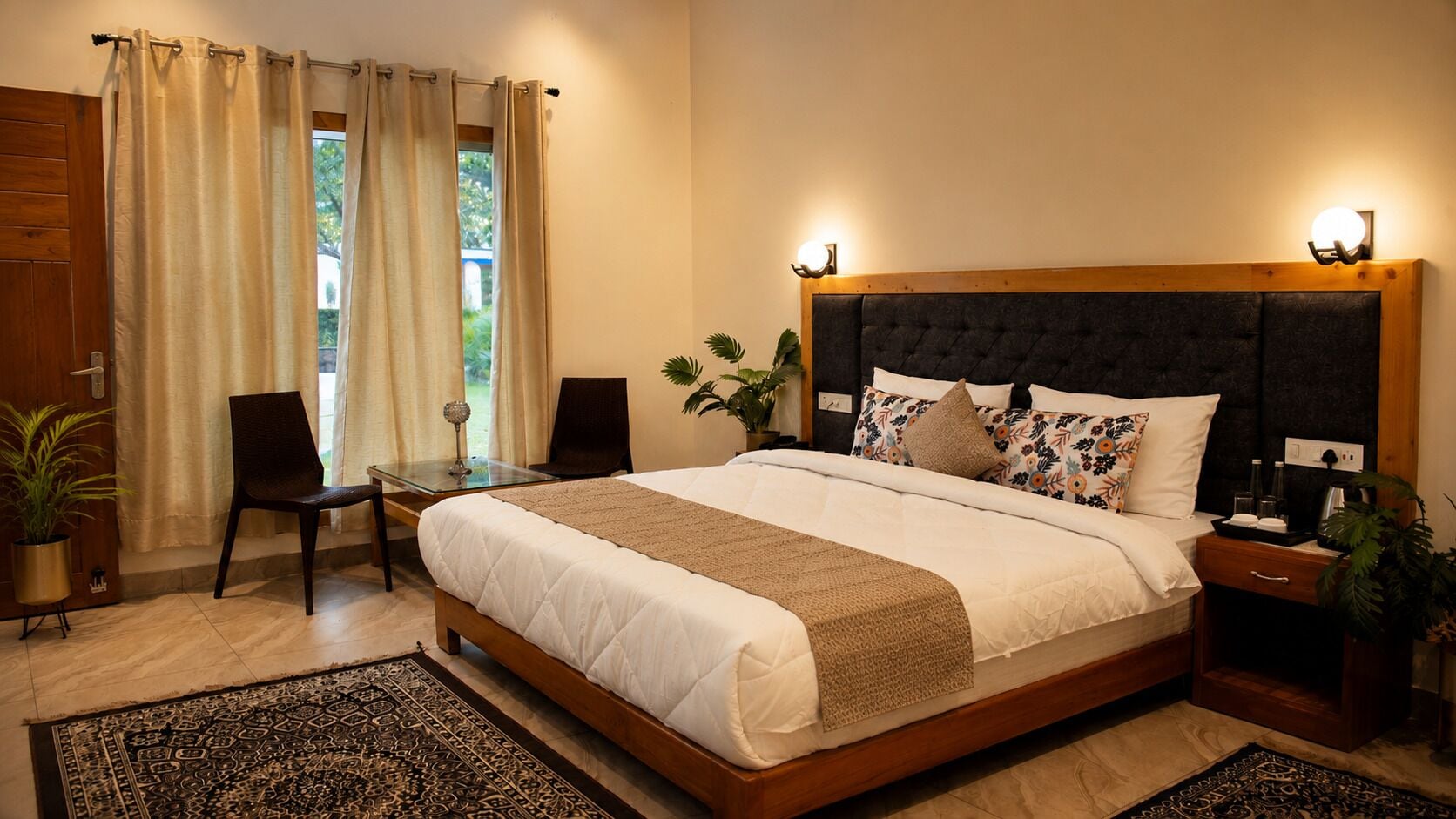 A bedroom with a large bed, bedside tables, a desk, chairs, and a view of the outdoors.