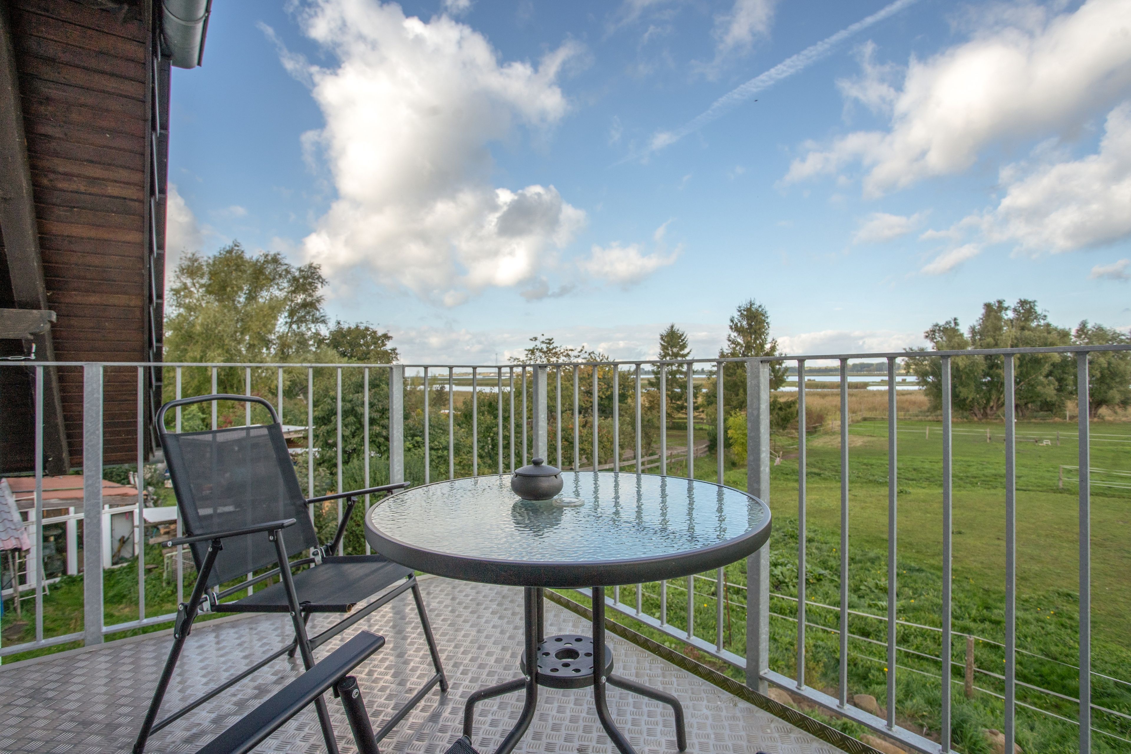 A balcony with a round glass table and two chairs, overlooking a grassy field.