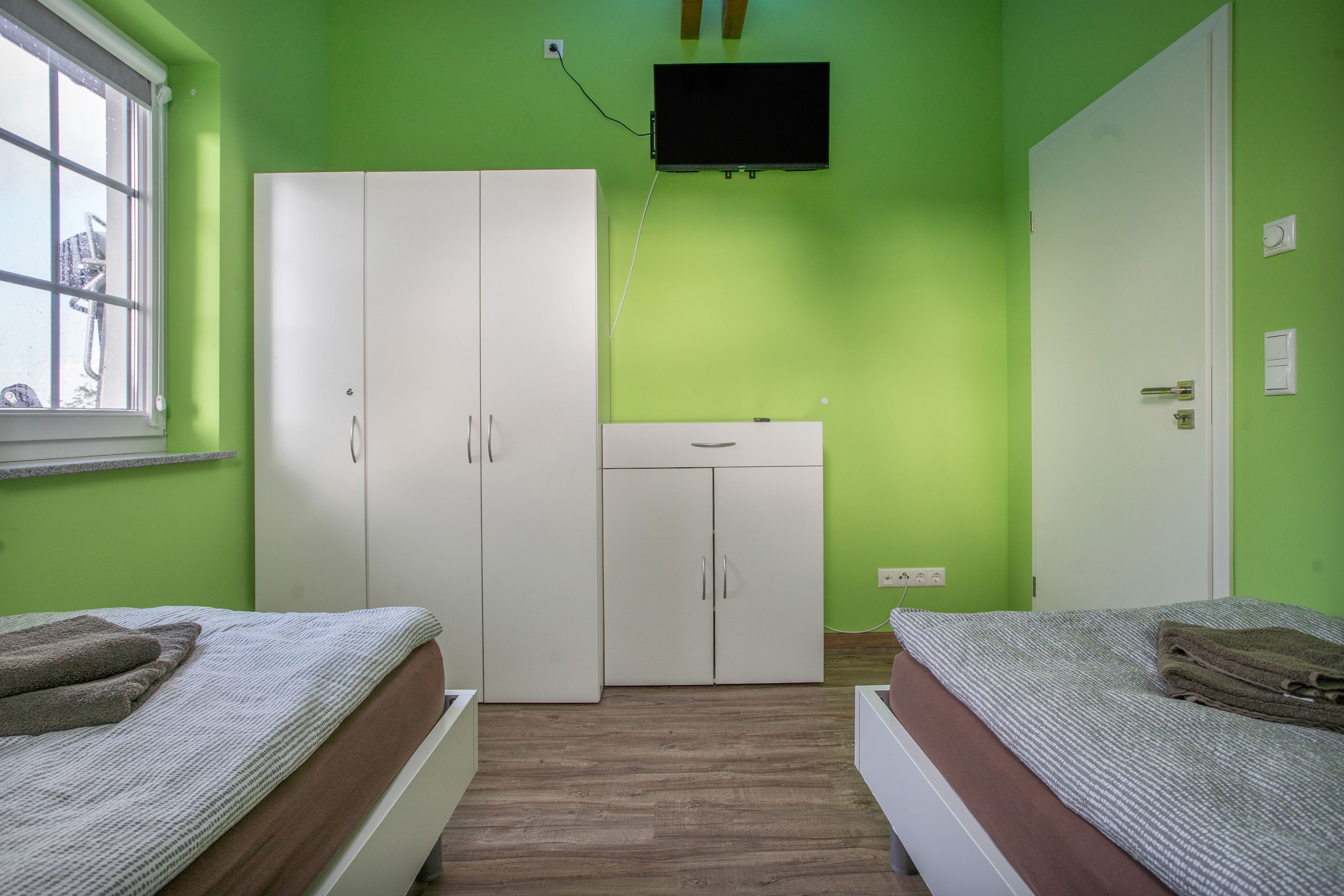 Ein Zimmer mit zwei Betten, einem Kleiderschrank, einem an der Wand montierten Fernseher und einem Fenster.