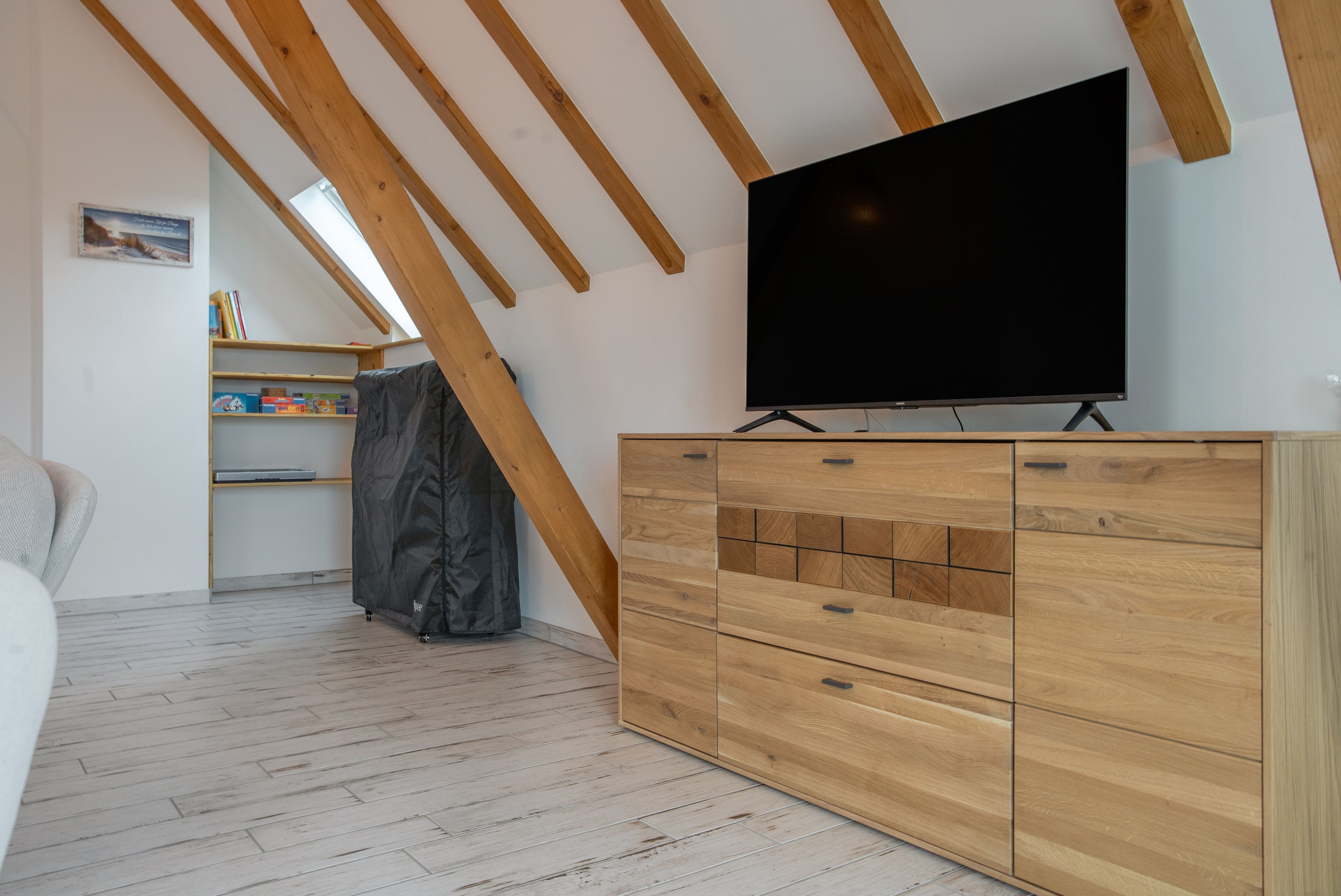 A room with a wooden ceiling, a flat-screen TV mounted on a wooden cabinet, a bookshelf, and a covered item on the floor.