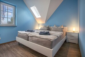 A bedroom with a sloped ceiling, a skylight, a bed with pillows and a blanket, bedside tables with lamps, and a window with a view of trees.