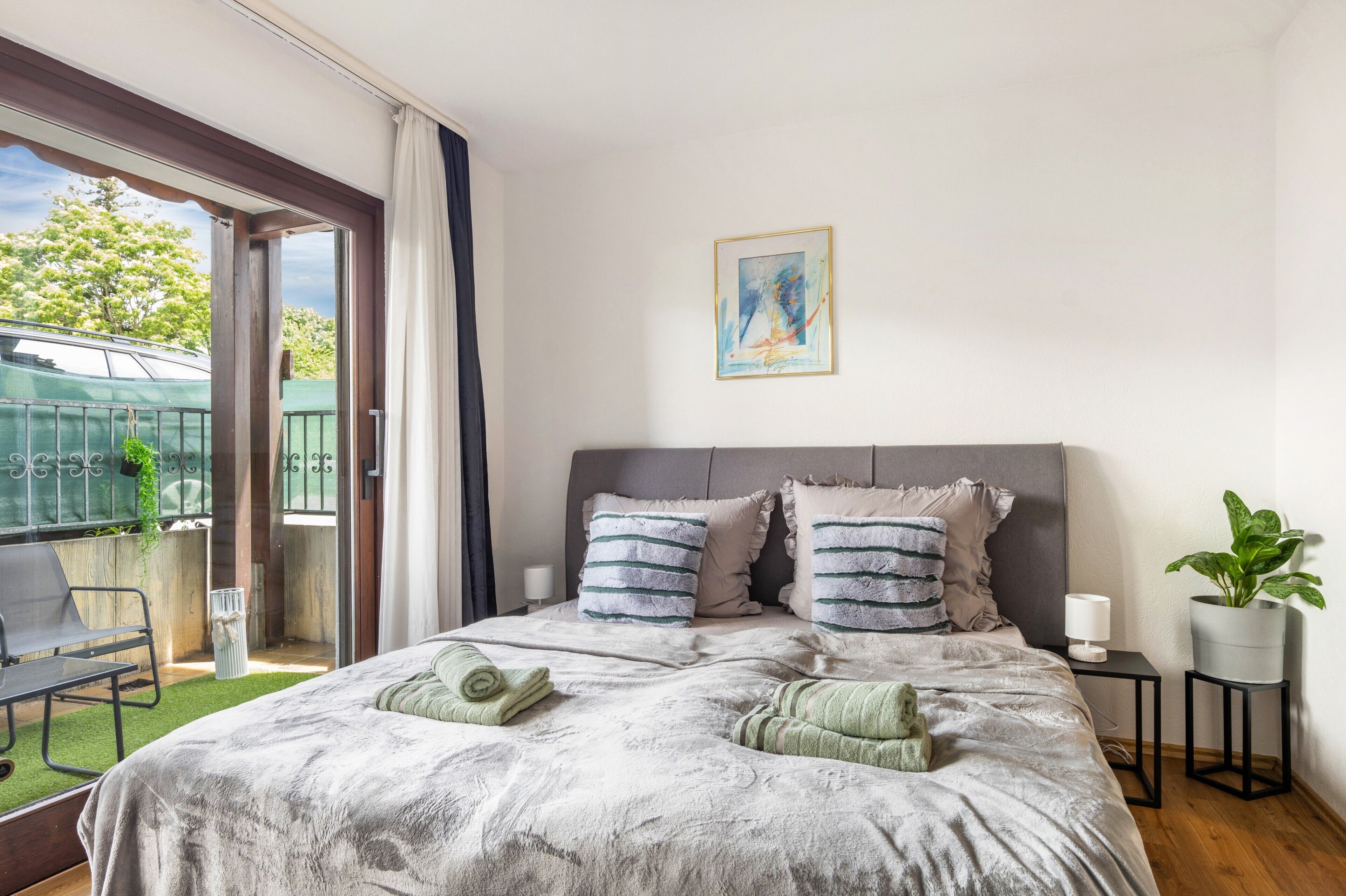 A bedroom with a bed, pillows, a bedside table, a potted plant, and a view of a balcony with a chair and a table.