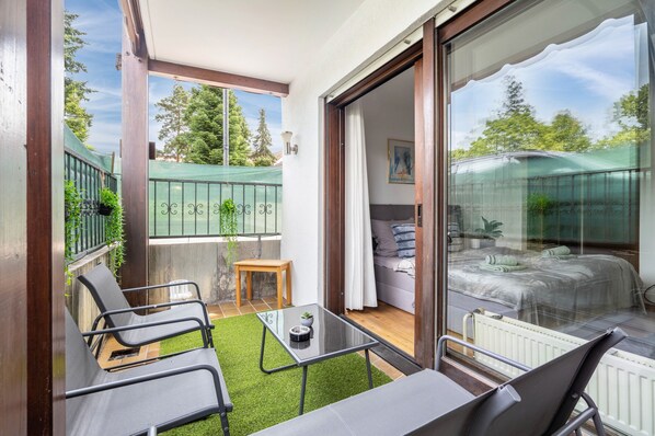 Un balcon avec une porte-fenêtre donnant sur une chambre, une petite table avec une plante et des sièges d’extérieur.