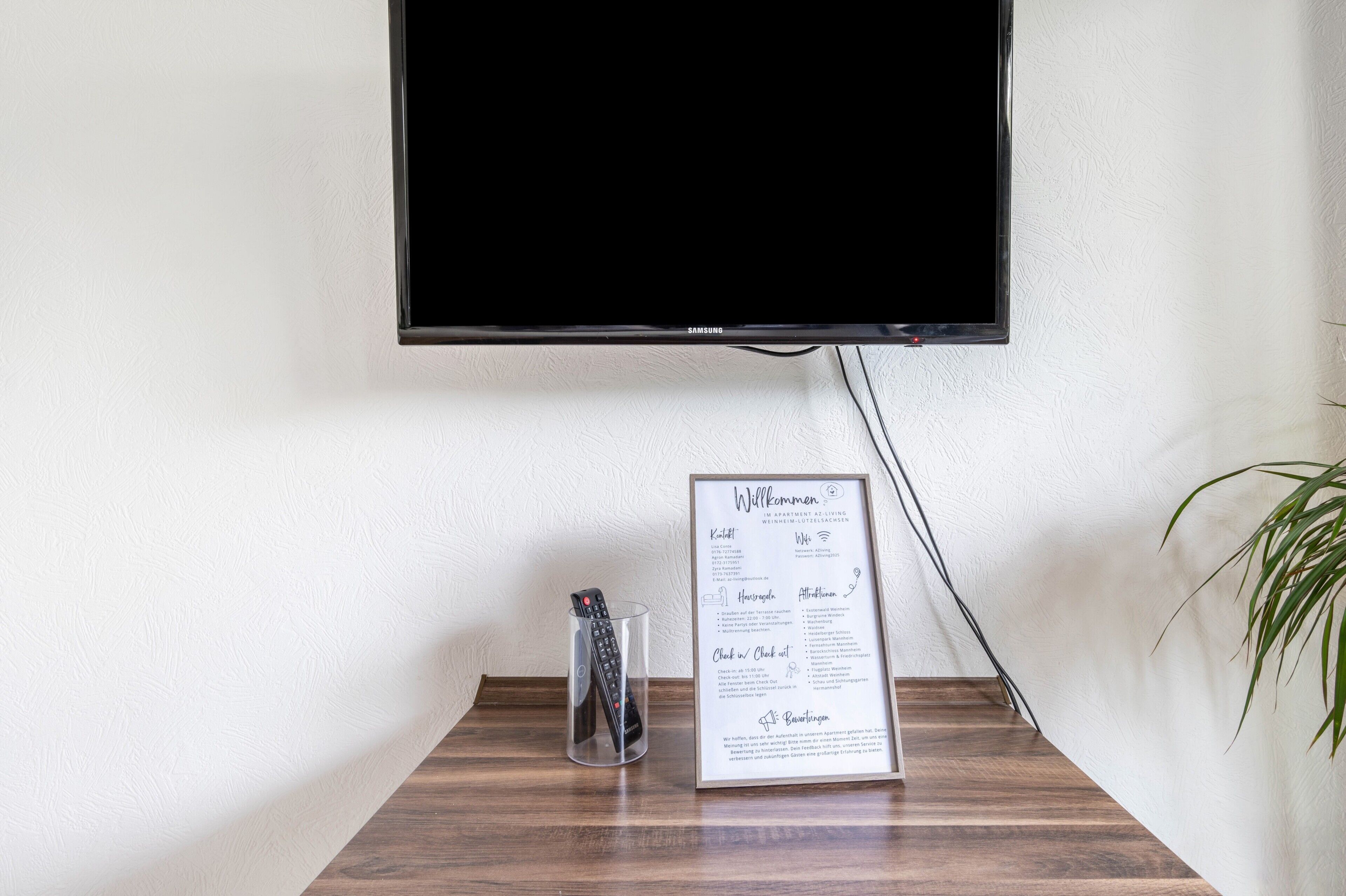 Un téléviseur à écran plat fixé au mur, un panneau d’accueil encadré avec les coordonnées, et un verre contenant une télécommande sur une table en bois.