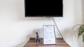 Un téléviseur à écran plat fixé au mur, un panneau d’accueil encadré avec les coordonnées, et un verre contenant une télécommande sur une table en bois.