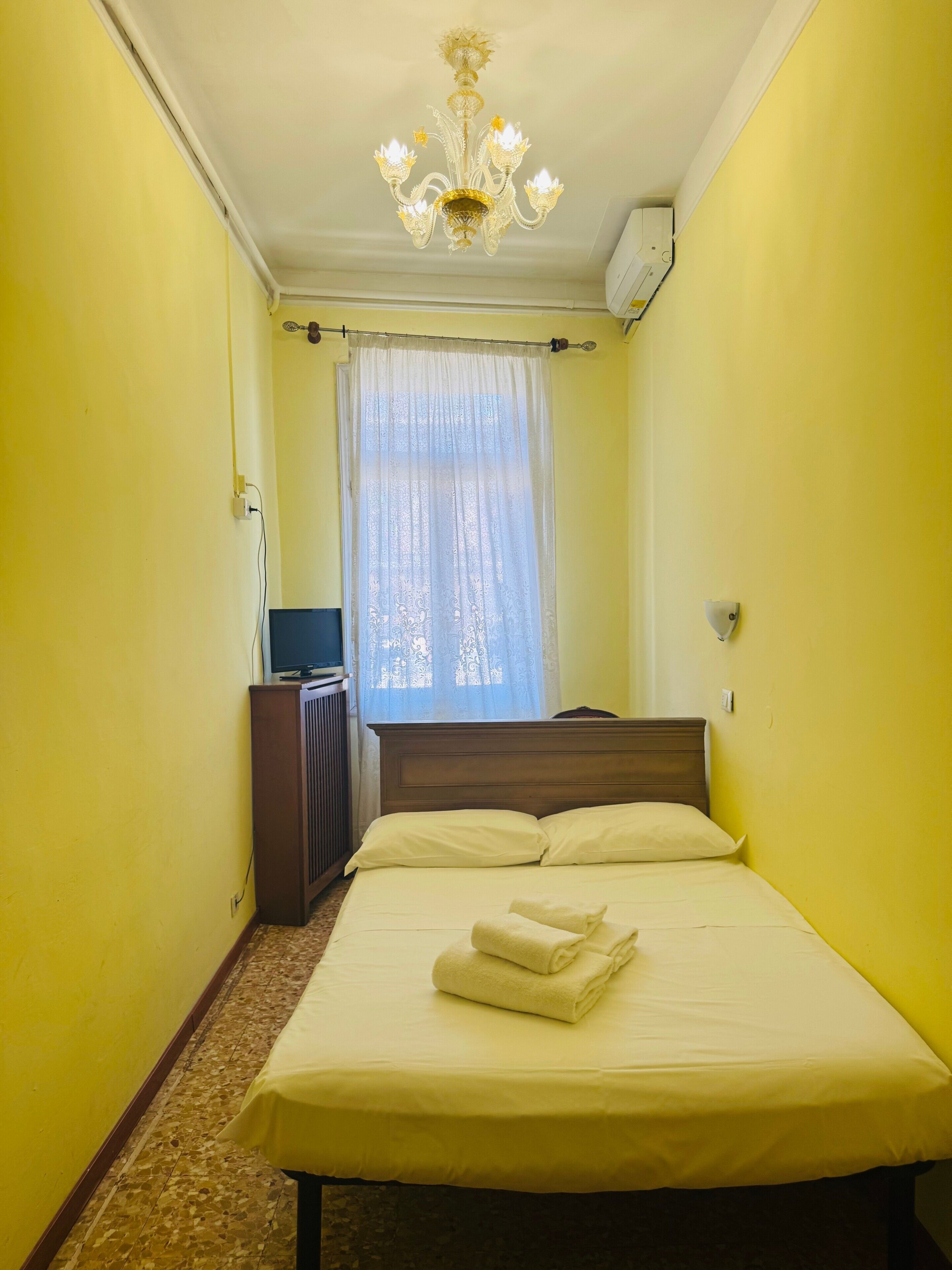 Une chambre avec un lit, une télévision, une fenêtre avec des rideaux et un lustre.