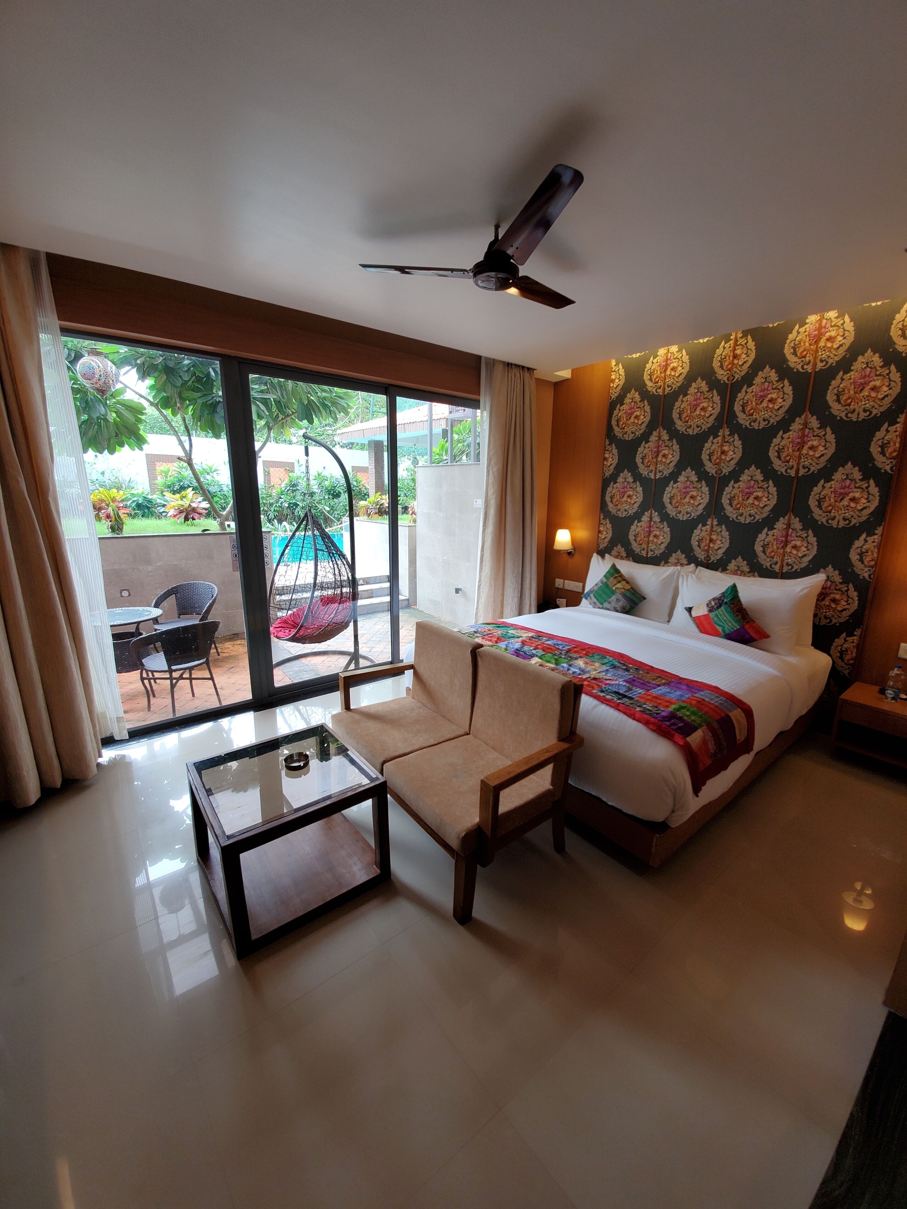 A hotel room with a bed, a chair, a table, a ceiling fan, and a view of the outside.