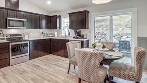 Eine moderne Küche mit dunklen Holzschränken, einer Edelstahl-Mikrowelle und einem eingebauten Backofen. Ein Essbereich mit gemusterten Stühlen und einem Tisch lädt zum Verweilen ein, und ein großes Fenster bietet einen Ausblick ins Freie.