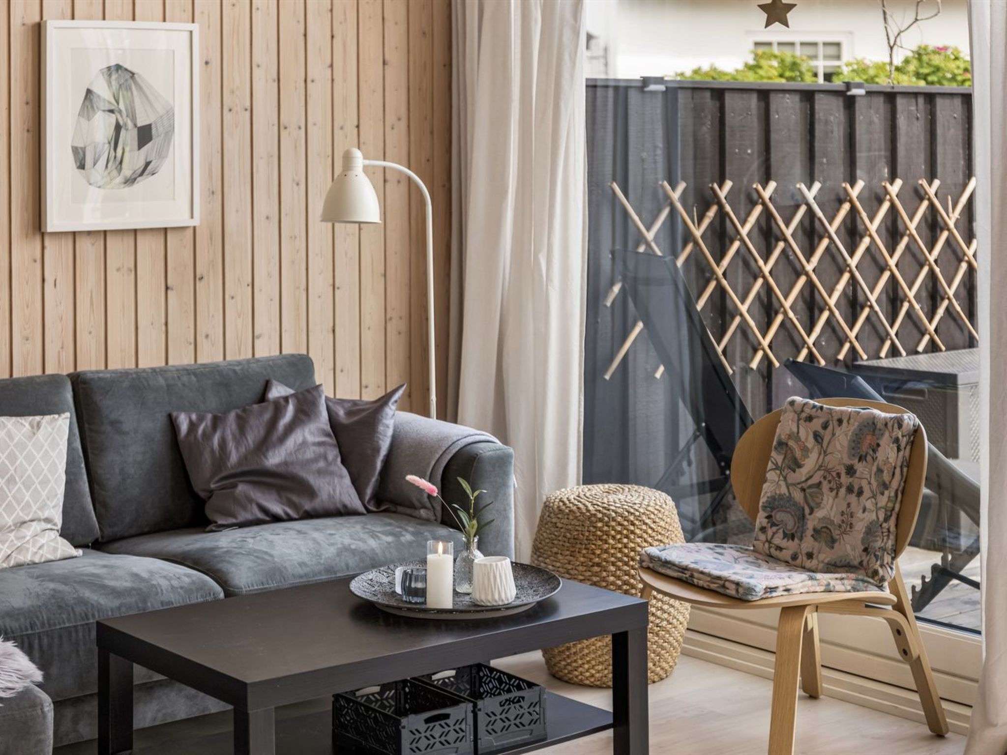 A modern living room with a grey sofa, a black coffee table, a wicker pouf, a wooden chair with a floral cushion, and a white floor lamp.