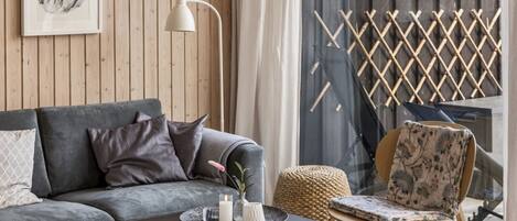 A modern living room with a grey sofa, a black coffee table, a wicker pouf, a wooden chair with a floral cushion, and a white floor lamp.