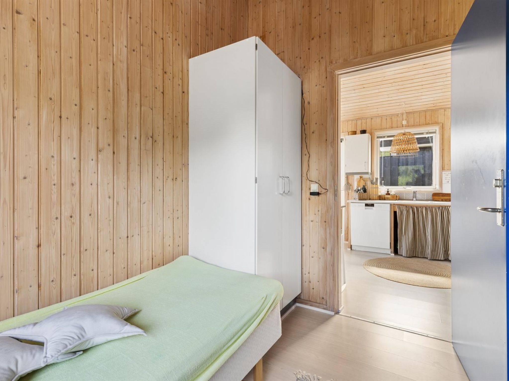 Une pièce comprenant un lit, une armoire blanche et un coin cuisine visible à travers une porte ouverte.