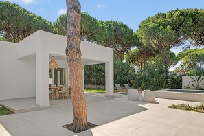 Un espace patio extérieur moderne, doté d’une structure blanche, de sièges et d’un grand arbre.