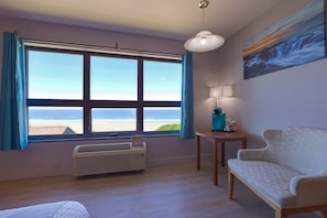 A room with a large window, a sofa, a table with a lamp, and a painting on the wall.