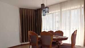 Un comedor con una mesa redonda y seis sillas, una araña, un ventanal con cortinas translúcidas y una cortina marrón a la izquierda.