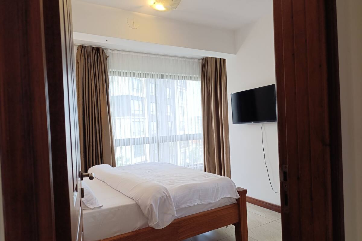 Un dormitorio con una cama de madera, un televisor instalado en la pared y una ventana con cortinas translúcidas.