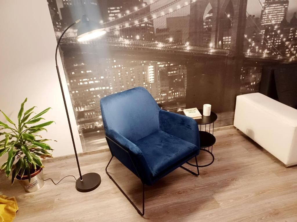 A room with a blue chair, a white sofa, a small table with a lamp, and a potted plant.