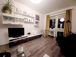 A living room with a flat-screen TV, a coffee table, a sofa, a dining table, and a chair. There are shelves with decorative items and a potted plant.
