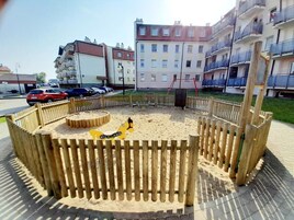 Un parque infantil con valla de madera, una excavadora de juguete amarilla y un arenero.