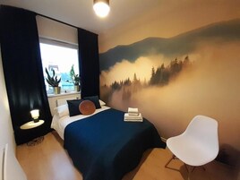 A bedroom with a bed, a white chair, a window, and a wall mural.